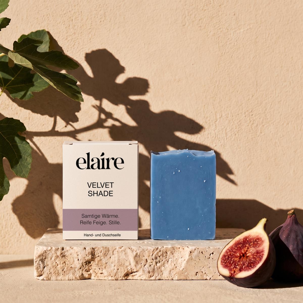 elaire Velvet Shade Seife mit Schachtel auf Travertin, daneben Feigen